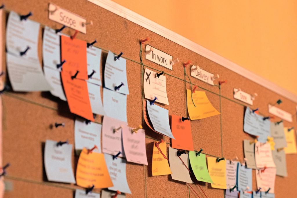 Close up of a Kanban board with sticky notes on it representing the scrum agile agile development methodology. There are columns labeled "Scope," "In work, and "Delivery"