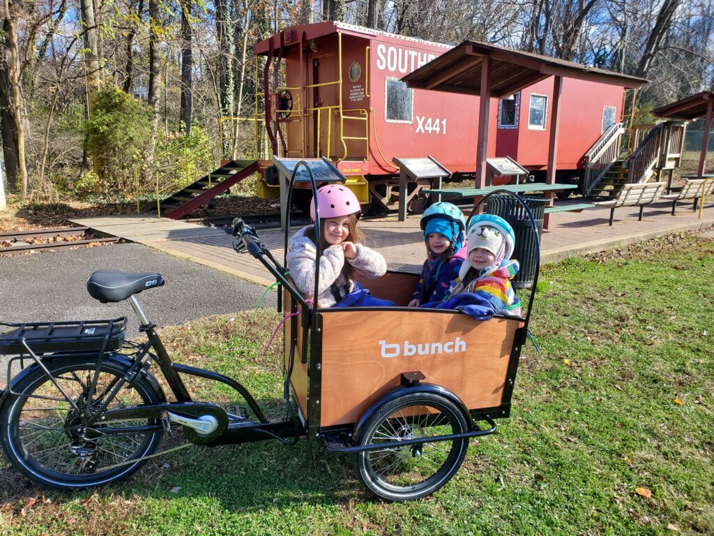 Image of Justin's daughters in their cargo bike. There is a cart attached to the front of the bike that the three daughters are seated in.