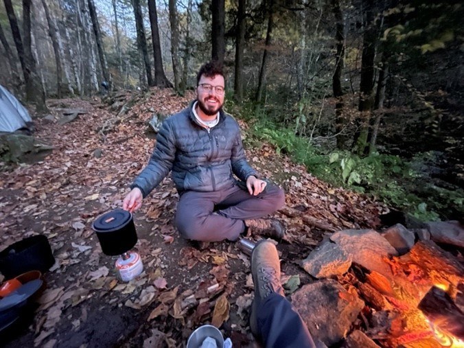 Adrien sitting on the ground in the woods next to campfire.