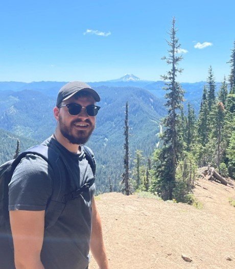 An image of Adrien on a trail in the mountains with trees and a large mountain in the background.