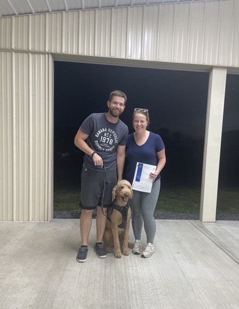 Megan and her husband standing with their dog holding up a therapy-dog certification.