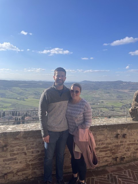 Megan and her husband posing against a stone wall with a backdrop of the hillside.